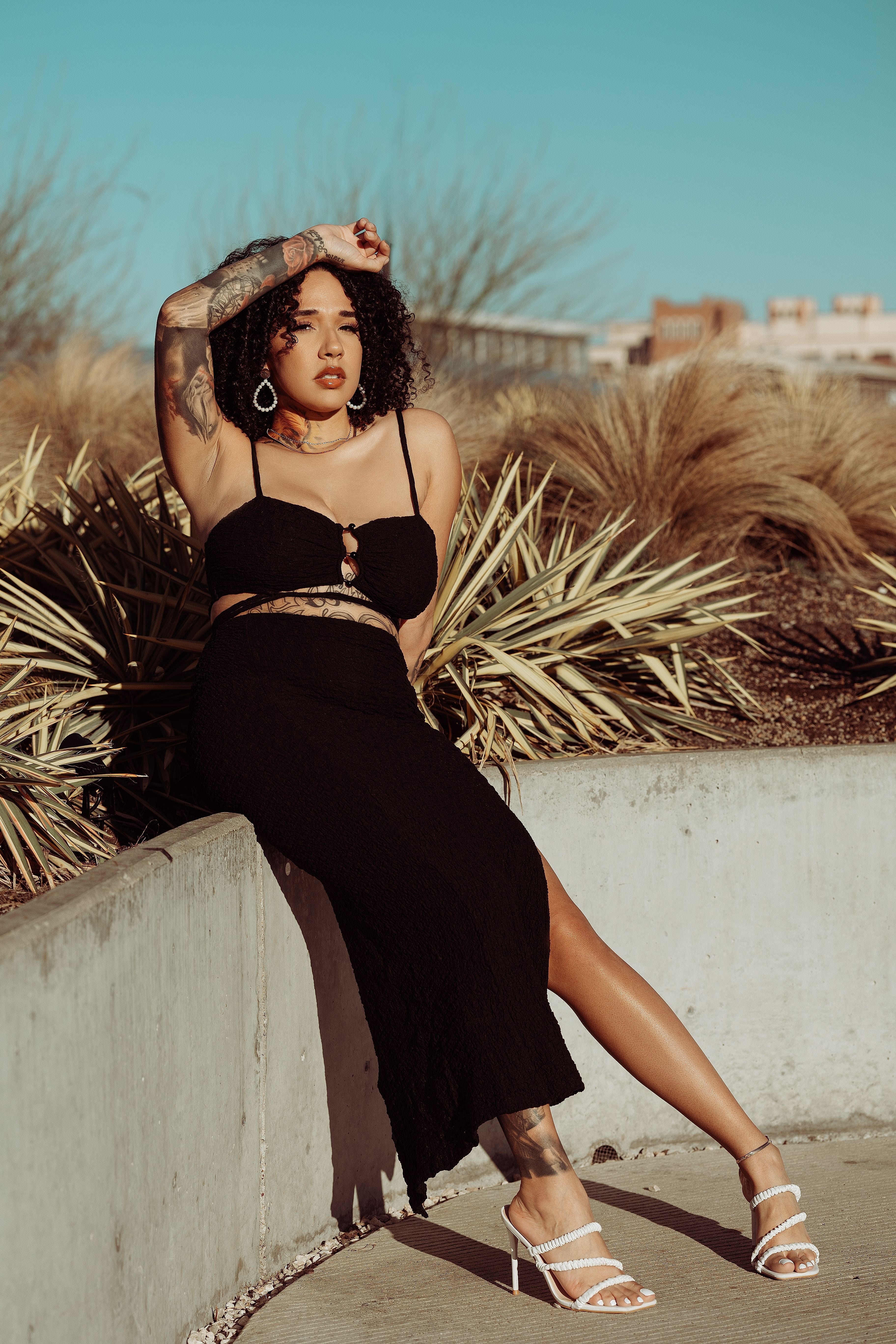 Woman in a black dress sitting outdoors with plants and a clear sky.